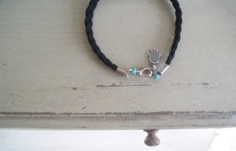 May include: A black braided cord bracelet with a silver charm of a hand and two turquoise beads.