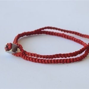 May include: A red braided cord bracelet with a silver square bead.