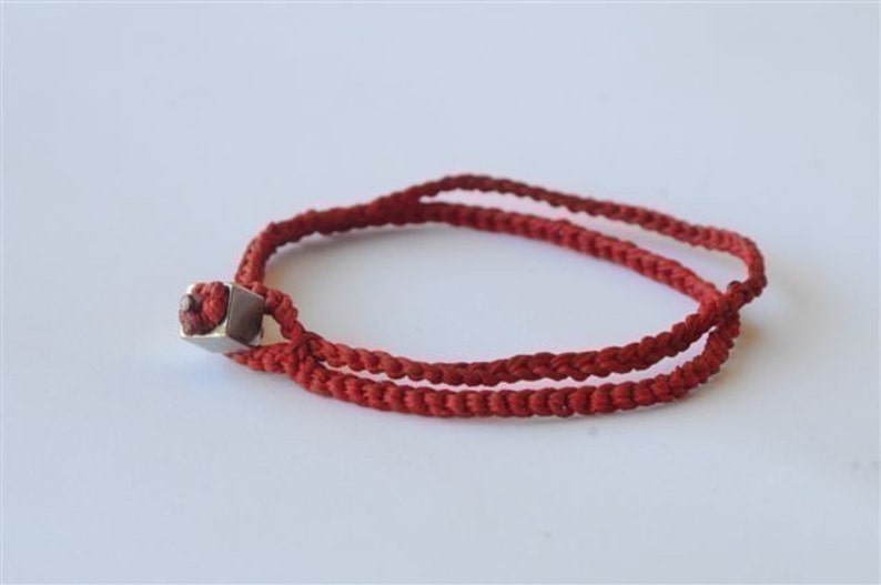 May include: A red braided cord bracelet with a silver square bead.