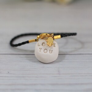 May include: Black braided bracelet with a gold clasp and a white ceramic charm that says "LOVE YOU" with a gold hamsa hand charm.