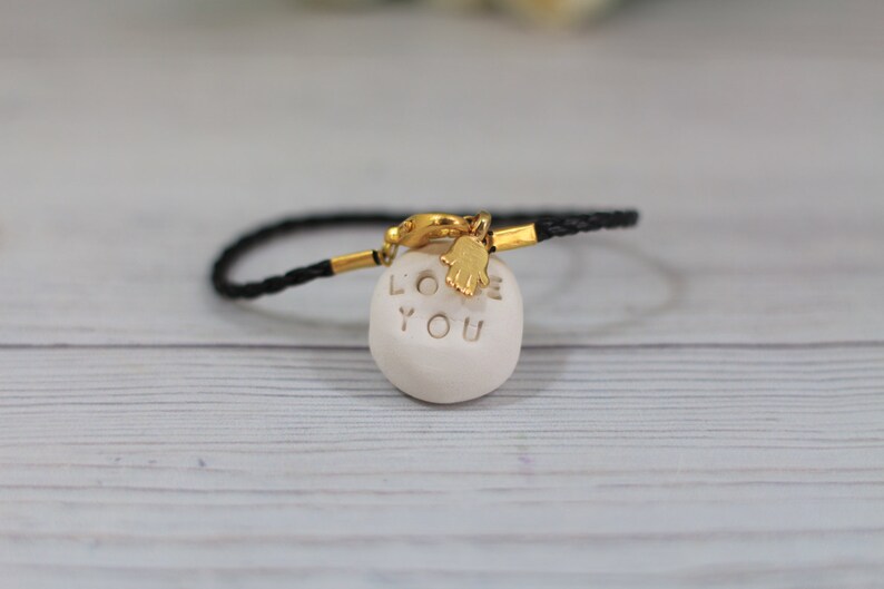 May include: Black braided bracelet with a gold clasp and a white ceramic charm that says "LOVE YOU" with a gold hamsa hand charm.