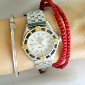 May include: A silver and gold wristwatch with a white face and black and blue accents. The watch has a silver metal band and is worn on a wrist with a red string bracelet.