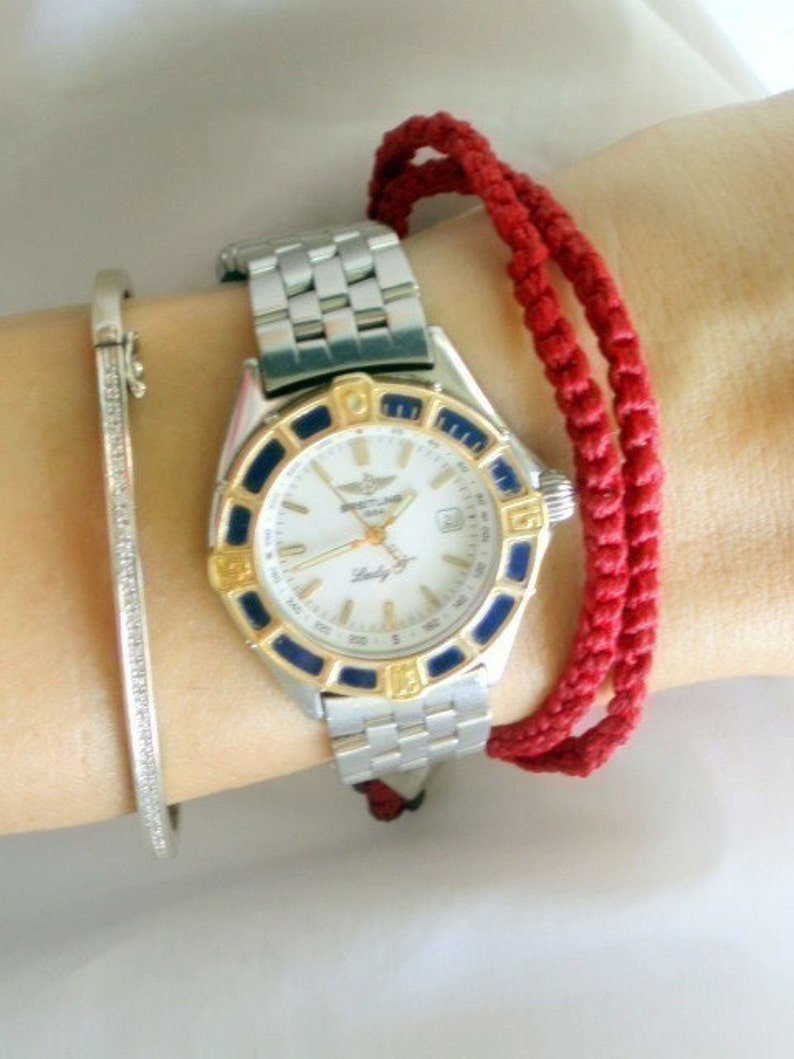May include: A silver and gold wristwatch with a white face and black and blue accents. The watch has a silver metal band and is worn on a wrist with a red string bracelet.