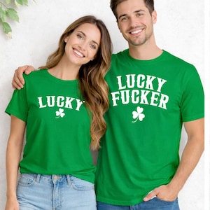 Op de afbeelding: Twee mensen dragen bijpassende groene t-shirts met witte tekst en een klavertje vier. Eén shirt zegt "LUCKY" en de andere zegt "LUCKY FUCKER". De shirts worden gecombineerd met een blauwe jeans.