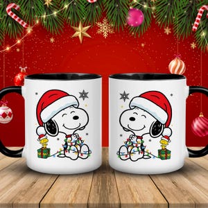 May include: Two white ceramic mugs with black handles and rims. Each mug features a cartoon Snoopy character wearing a Santa hat and wrapped in Christmas lights. A small yellow bird is on a gift box.