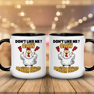 Puede incluir: Dos tazas de cerámica blancas con asas y bordes negros. Cada taza presenta un dibujo de un pollo con el texto "DON'T LIKE ME? CAW F PROBLEM SOLVED" en negro, rojo y amarillo. Las tazas están sobre una superficie de madera.