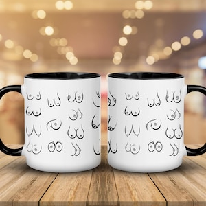 Puede incluir: Dos tazas de café blancas con asas y bordes negros. Las tazas están decoradas con un patrón repetido de dibujos lineales negros de senos. Las tazas están sobre una superficie de madera.