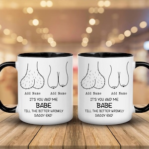 Op de afbeelding: Twee witte keramische mokken met zwarte handvatten en randen. Elke mok heeft een humoristische illustratie van de mannelijke anatomie en de tekst "IT'S YOU AND ME BABE TILL THE BITTER WRINKLY SAGGY END".