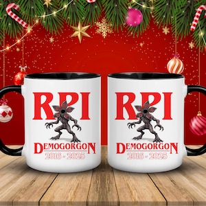 May include: Two white ceramic mugs with black handles and rims. Each mug features the word "RPI" in red, a Demogorgon illustration, and the text "DEMOGORGON 2016-2025" in red. The mugs are on a wooden surface with Christmas decorations in the background.