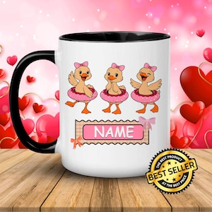May include: White ceramic mug with a black handle and interior, featuring three cartoon ducklings wearing pink bows and inner tubes. A banner with the word "NAME" is below the ducks. A "Best Seller" sticker is on the bottom right.