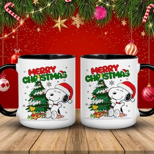 Könnte beinhalten: Zwei weiße Keramikbecher mit schwarzen Griffen und Rändern. Jeder Becher zeigt eine Snoopy-Grafik mit Weihnachtsmütze, einen Weihnachtsbaum und die Worte "MERRY CHRISTMAS" in Grün und Rot. Die Becher stehen auf einer Holzoberfläche.