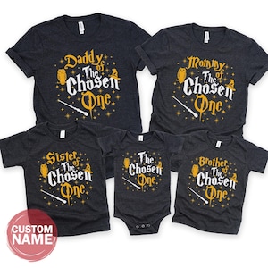 May include: A set of matching dark gray t-shirts and a onesie with the text "Daddy of The Chosen One", "Mommy of The Chosen One", "Sister of The Chosen One", "Brother of The Chosen One", and "The Chosen One". Each shirt features a wand, stars, and an owl.