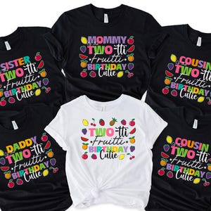 May include: A collection of black and white t-shirts with colorful text and fruit graphics. The shirts feature phrases like "Sister of the Two-tti Fruitti Birthday Cutie" and "Mommy of the Two-tti Fruitti Birthday Cutie."