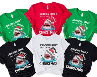 May include: A collection of Christmas-themed t-shirts in various colors, including green, red, black, and white. Each shirt features a shark wearing a Santa hat, with the text "JOHNSON FAMILY Have a Holly JAWLLY CHRISTMAS".