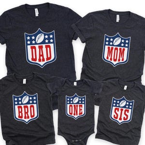 May include: A collection of dark gray t-shirts and a onesie, each featuring a shield design with a football and stars. The shirts display the words "DAD", "MOM", "BRO", and "SIS", while the onesie says "ONE".