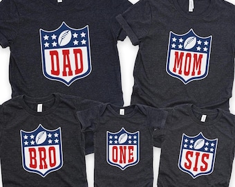 May include: A collection of dark gray t-shirts and a onesie, each featuring a shield design with a football and stars. The shirts display the words "DAD", "MOM", "BRO", and "SIS", while the onesie says "ONE".