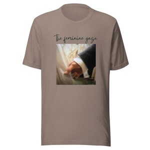 Op de afbeelding: Een lichtbruin T-shirt met een kleine vierkante afbeelding van een hand die stof aanraakt, met de tekst "The feminine gaze" in cursiefschrift erboven. Het shirt is gemaakt van een zacht materiaal.