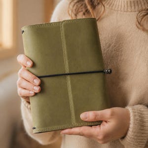 Könnte beinhalten: Ein salbeigrüner Einband für ein Notizbuch aus Leder mit matter Oberfläche und sichtbaren Nähten an den Rändern. Der Einband wird durch ein dünnes schwarzes Gummiband gesichert, das horizontal um die Mitte verläuft und von einer kleinen Metallperle gehalten wird.