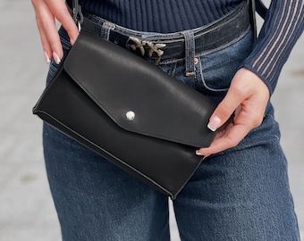Könnte beinhalten: Eine schwarze Leder-Umhängetasche mit Klappenverschluss und silbernem Druckknopf. Die Tasche wird von einem schwarzen Riemen gehalten und über der Schulter getragen. Die Tasche ist rechteckig und scheint leer zu sein.