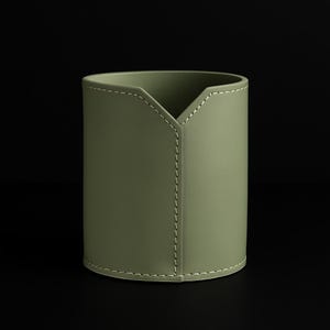 May include: An olive green cylindrical desk tidy with a V-shaped top and white stitching detail. The organiser is made from a smooth material and has a minimalist design. Suitable for storing pens, pencils, and other stationery items.