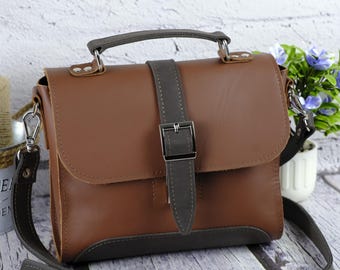 Könnte beinhalten: Eine braune Ledertasche mit grauem Riemen und Griff. Die Tasche hat eine silberne Schnalle und einen abnehmbaren Schulterriemen. Die Tasche ist etwa 25 cm breit und 20 cm hoch.