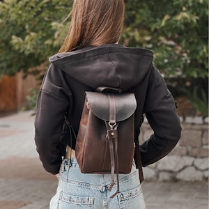 Può includere: Uno zaino in pelle marrone con chiusura a patta e spallacci regolabili. Lo zaino è indossato sulla schiena di una persona che indossa una felpa con cappuccio nera e jeans blu. La borsa ha un design semplice e classico.
