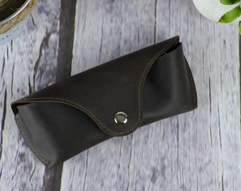 May include: A dark brown leather glasses case with a press stud closure. The case is rectangular with rounded edges and stitched detailing. It is designed to protect spectacles or sunglasses.
