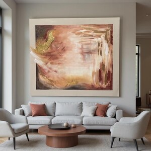 Puede incluir: Gran pintura abstracta con borde beige, con tonos cálidos de oro, marrón y melocotón. La obra de arte cuelga sobre un sofá gris claro con cojines color óxido. Una mesa de centro redonda de madera y dos sillones completan el salón.