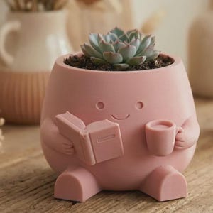 Puede incluir: Una maceta rosa con forma de personaje, sosteniendo un libro y una taza, llena de tierra y una planta suculenta. El diseño presenta una cara sonriente y rasgos redondeados. Un complemento encantador para la decoración del hogar.