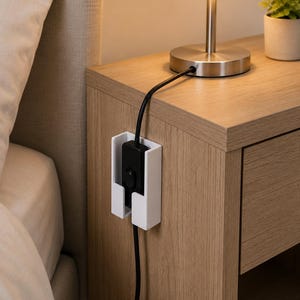 May include: A white rectangular wall-mounted holder attached to the side of a wooden nightstand. The holder contains a black electrical power cord with an inline switch, which runs vertically through the centre of the white plastic bracket.