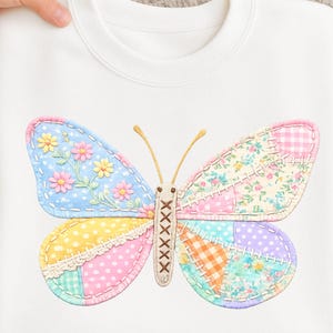 Op de afbeelding: Witte sweatshirt met een kleurrijk vlinderontwerp. De vlindervleugels zijn gemaakt van stoffen met patronen in pasteltinten, waaronder bloemen-, stippen- en vichy-patronen. Het lichaam van de vlinder is bruin met gestikte details.