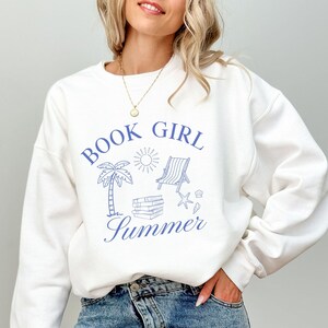 May include: White sweatshirt with blue graphic text and illustrations. The text reads "BOOK GIRL SUMMER" with drawings of a palm tree, sun, beach chair, books, and seashells. The sweatshirt is a casual, comfortable style.