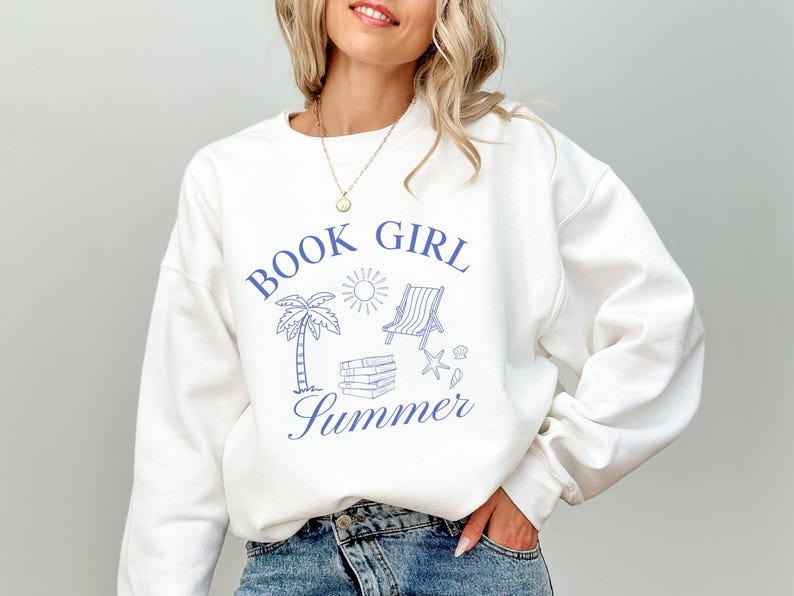 May include: White sweatshirt with blue graphic text and illustrations. The text reads "BOOK GIRL SUMMER" with drawings of a palm tree, sun, beach chair, books, and seashells. The sweatshirt is a casual, comfortable style.
