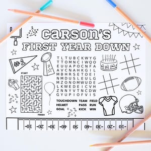 May include: A black and white coloring page with the text "Carson's First Year Down" and football-themed illustrations. Includes a maze, football, helmet, and word search. Colored pencils with blue, red, and yellow tips are scattered around the page.