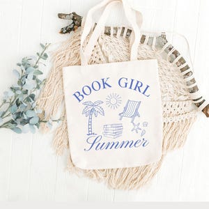 May include: A cream-colored tote bag with blue text and illustrations. The text reads "BOOK GIRL Summer" with images of a palm tree, sun, beach chair, books, and seashells. The bag has two handles and is displayed on a white surface.