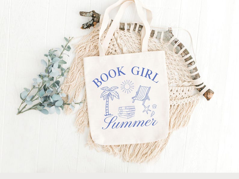 May include: A cream-colored tote bag with blue text and illustrations. The text reads "BOOK GIRL Summer" with images of a palm tree, sun, beach chair, books, and seashells. The bag has two handles and is displayed on a white surface.