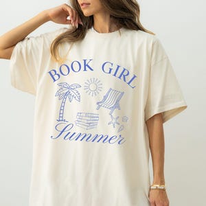 May include: Cream-colored t-shirt with a blue graphic that reads "BOOK GIRL Summer." The design includes a palm tree, sun, beach chair, books, and seashells. The shirt has short sleeves and a relaxed fit.