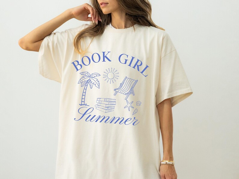 May include: Cream-colored t-shirt with a blue graphic that reads "BOOK GIRL Summer." The design includes a palm tree, sun, beach chair, books, and seashells. The shirt has short sleeves and a relaxed fit.
