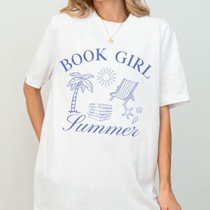 May include: White t-shirt with a blue graphic that reads "Book Girl Summer." The design includes a palm tree, sun, beach chair, books, and starfish. A casual, comfortable tee for book lovers.