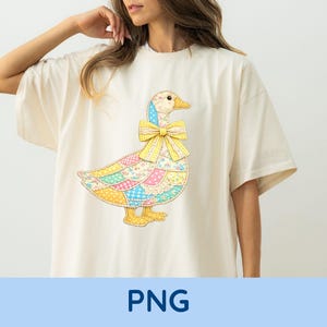 Könnte beinhalten: Cremefarbenes T-Shirt mit einem bunten Patchwork-Gans-Design und einer gelben Schleife. Die Gans hat einen blauen Hut und gelbe Füße. Das Shirt hat kurze Ärmel und eine lockere Passform.