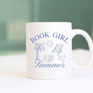 May include: White ceramic mug with a blue design featuring the words "BOOK GIRL Summer" along with illustrations of a palm tree, sun, beach chair, books, and seashells. The mug has a curved handle.