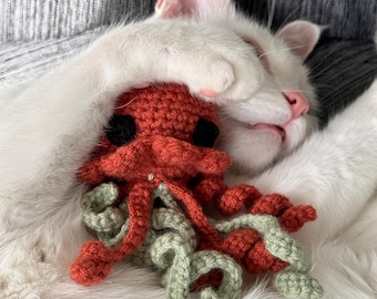 May include: A white cat with closed eyes is resting, covering its face with a paw. A handmade, crocheted octopus in shades of orange and green rests on the cat's chest. The octopus has black eyes.