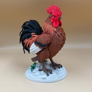 May include: A three-dimensional rooster figurine featuring a bright red comb and wattle, brown body feathers, black tail feathers, and white wing accents. The bird stands on a textured white base with small green grass details.