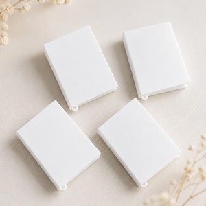 May include: Four rectangular white book-shaped charms arranged in a grid. Each charm features a smooth, solid white surface and a small, integrated loop at the bottom edge, designed for attachment to jewelry or other decorative items.