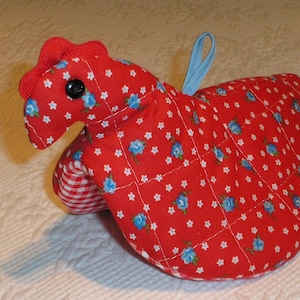 May include: A quilted red chicken-shaped oven mitt with a blue loop for hanging. The mitt features a floral pattern with blue flowers and white stars. The comb is red, and the eye is a black button.