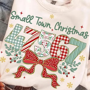 Puede incluir: Sudadera blanca con la frase "Small Town Christmas" en verde. Los números "47567" están diseñados con varios patrones, incluyendo cuadros, rayas y estampados florales. Un lazo rojo con lunares en la parte inferior, con copos de nieve y bayas.