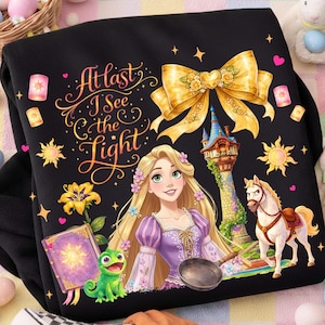 May include: Black sweatshirt featuring a colorful graphic of a princess, a horse, and a tower. The design includes the text "At last I See the Light" in gold script, along with floral and star accents. A large gold bow is at the top.