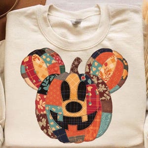 May include: Cream-colored sweatshirt featuring a patchwork Mickey Mouse pumpkin design. The pumpkin has a smiling face and is made of various fabric patterns in orange, red, blue, and brown. The sweatshirt is a crew neck style.