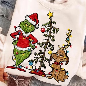 Puede incluir: Sudadera blanca con un personaje de Grinch con gorro de Papá Noel y abrigo rojo, un árbol de Navidad y un perro marrón con cuernos. El Grinch es verde con un atuendo rojo y blanco. El perro es marrón con un collar rojo.