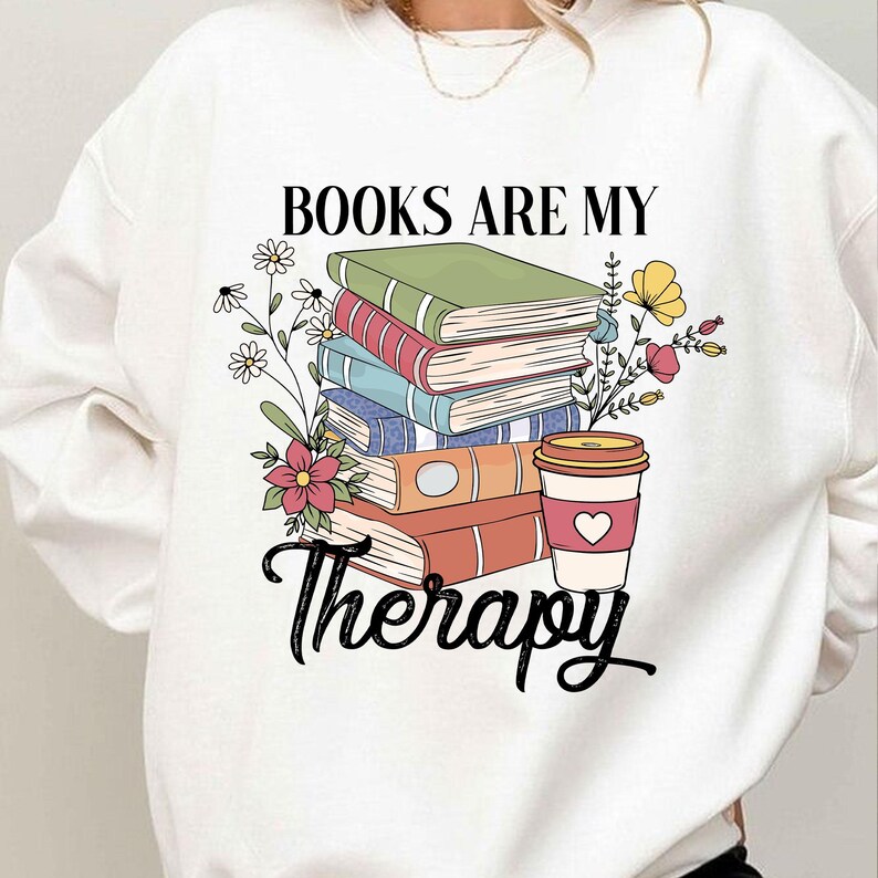 May include: White sweatshirt with a graphic design of a stack of books and a coffee cup. The text "Books are my therapy" is printed on the sweatshirt.